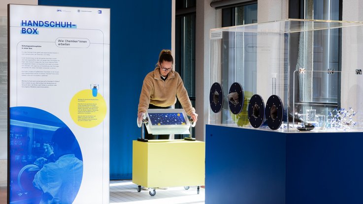 VISUELL Szenografie: Wissenschaftsausstellung „Was kann Katalyse?“ für den Sonderforschungsbereich SFB 1333 der Universität Stuttgart VISUELL Szenografie: Wissenschaftsausstellung: Eine Besucherin bedient eins der zwei interaktiven Exponate in der Ausstellung „Was kann Katalyse?“, daneben befindet sich eine Informationstafel mit Text und Bild