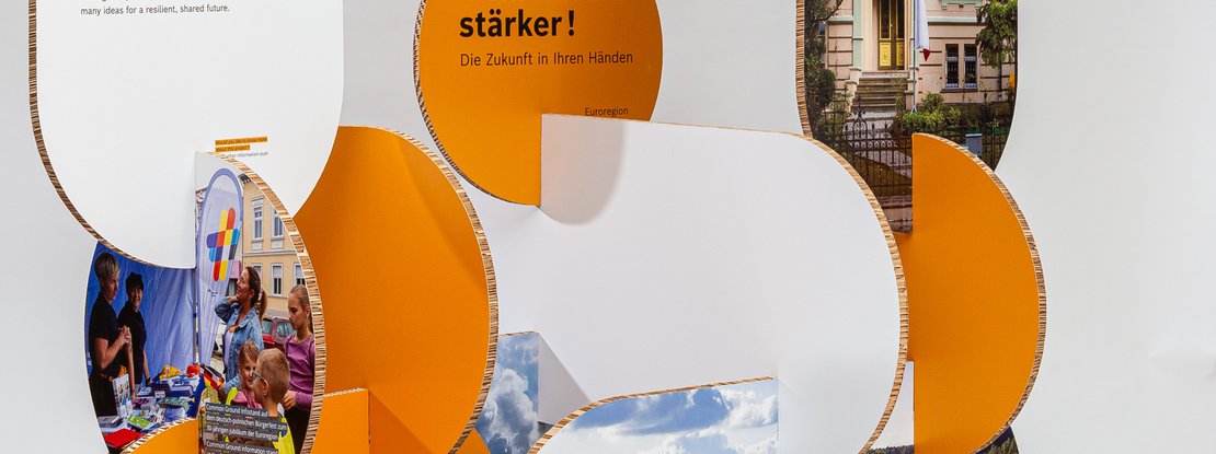 VISUELL Szenografie: Wanderausstellung „Common Grounds – Über Grenzen mitgestalten“ für die Robert Bosch Stiftung GmbH VISUELL Szenografie: Wanderausstellung Robert Bosch Stiftung GmbH: Detailansicht einer organisch geformten Ausstellungsstation aus orangefarbenen und weißen Wabenplatten; integrierte Bildtafeln zeigen Eventszenen und Landschaften, darüber ein Schriftzug der mit „…stärker! Die Zukunft in Ihren Händen“ endet.