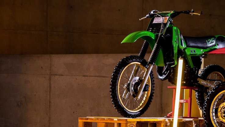 VISUELL Szenografie: Ausstellung für Mobilitäts- und Sportverband VISUELL Szenografie: Ausstellung: Ein grünes Motorrad wird auf Paletten und mit Lichtstelen in Szene gesetzt; Betonwand und Bodenmarkierungen verstärken das industrielle Ambiente.