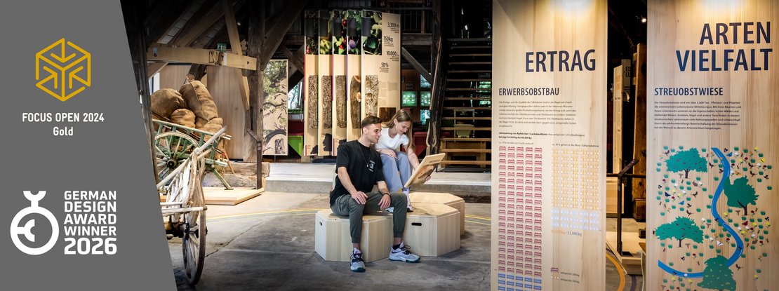 VISUELL Szenografie: Konzeption und Realisierung des Obstbaumuseums in Glems VISUELL Szenografie: Obstbaumuseum: Zwei Besucher sitzen in der Mitte der Ausstellung und betrachten eine Informationstafel in ihren Händen. Links neben dem Bild stehen Logos von Auszeichnungen, die das Projekt erhalten hat: Focus Open 2024 in Gold und German Design Award Winner 2026