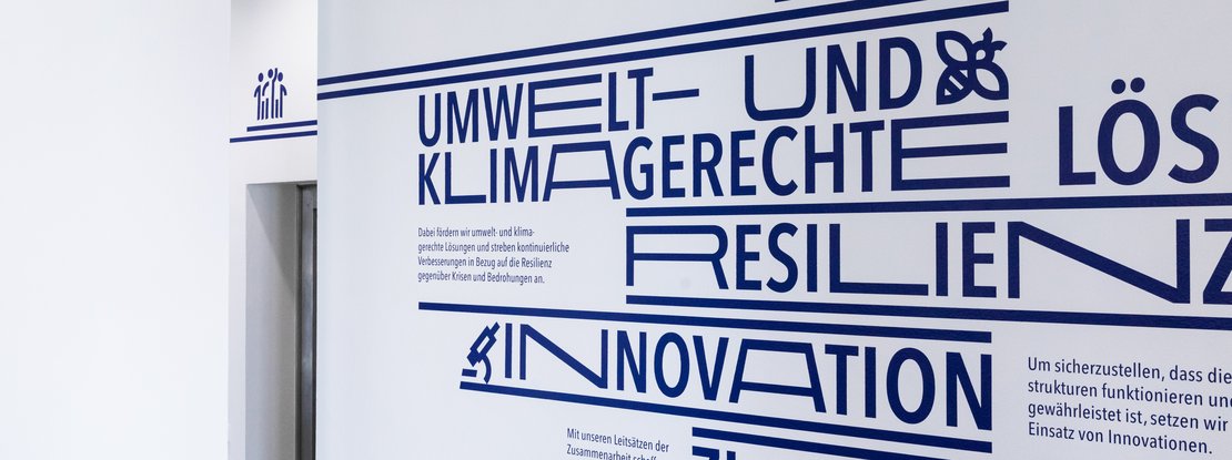 VISUELL Kommunikationsdesign: Wandgrafiken für die Landeshauptstadt Stuttgart VISUELL Kommunikationsdesign: Wandgrafiken Landeshauptstadt Stuttgart: Großformatige Wandgrafik mit typografisch hervorgehobenen Begriffen wie Umwelt- und klimagerechte Lösungen, Resilienz und Innovation in tiefem Dunkelblau auf weißer Wand.