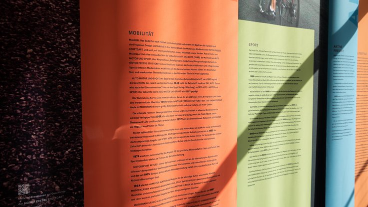 VISUELL Szenografie: Ausstellungsrundgang: Detailaufnahme Stoffbanner orange, grün und blau - Aufschrift: Mobilität, Sport, Aktive Freizeit