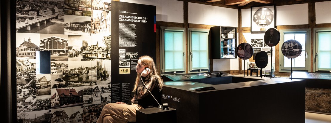VISUELL Szenografie: Konzeption und Umsetzung der Dauerausstellung Stadtmuseum Hornmoldhaus VISUELL Szenografie: Dauerausstellung Hornmoldhaus: Historischer Ausstellungsraum mit sichtbaren Holzbalken und zentralem schwarzem Ausstellungstisch. Eine Besucherin nutzt eine Hörstation mit Kopfhörer. Großformatige Texttafel „Zusammenschluss – Zusammenwachsen“ und historische Fotocollage strukturieren die chronologische Stadtgeschichte.