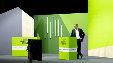 VISUELL Szenografie: Bühnenbild und Gesamtinszenierung: Mann steht hinter grünem Rednerpult vor grünem Bühnenbild mit vereinzelten Pflanzen