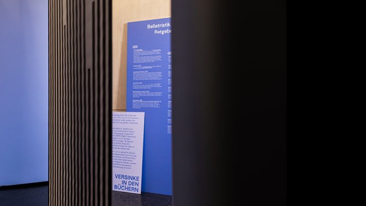 VISUELL Szenografie: Ausstellungsrundgang: zwei blaue Texttafeln – Aufschrift: Versinke in den Büchern